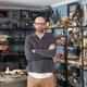 Producer Brian Volk-Weiss wears a grey long sleeve top and beige pants. He stands with his arms crossed surrounded by a large number of shells with a ridiculously large amount of toys on them.